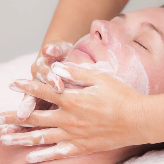 A person receiving a gentle facial massage, with eyes closed, and a creamy white mask applied. Hands are massaging the face, creating a relaxing atmosphere.