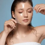 A woman with closed eyes peels a clear facial mask off her face. She has wet hair, a towel wrapped around her, against a light blue background.