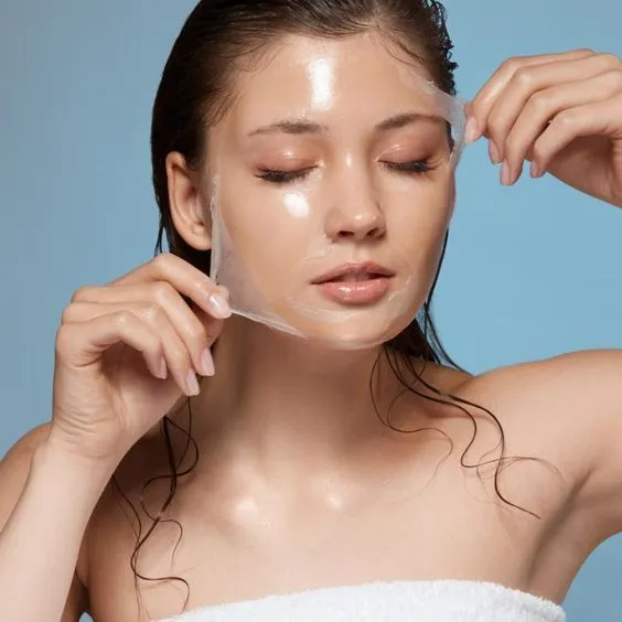 A woman with closed eyes gently removes a clear facial mask, revealing smooth skin. Her expression is relaxed. Background is a light blue.