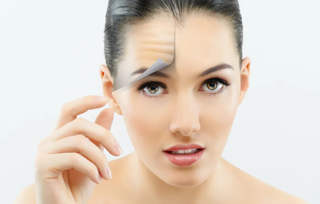 A woman with smooth skin peels back a photo of her forehead, revealing wrinkles underneath. She appears curious against a plain background.