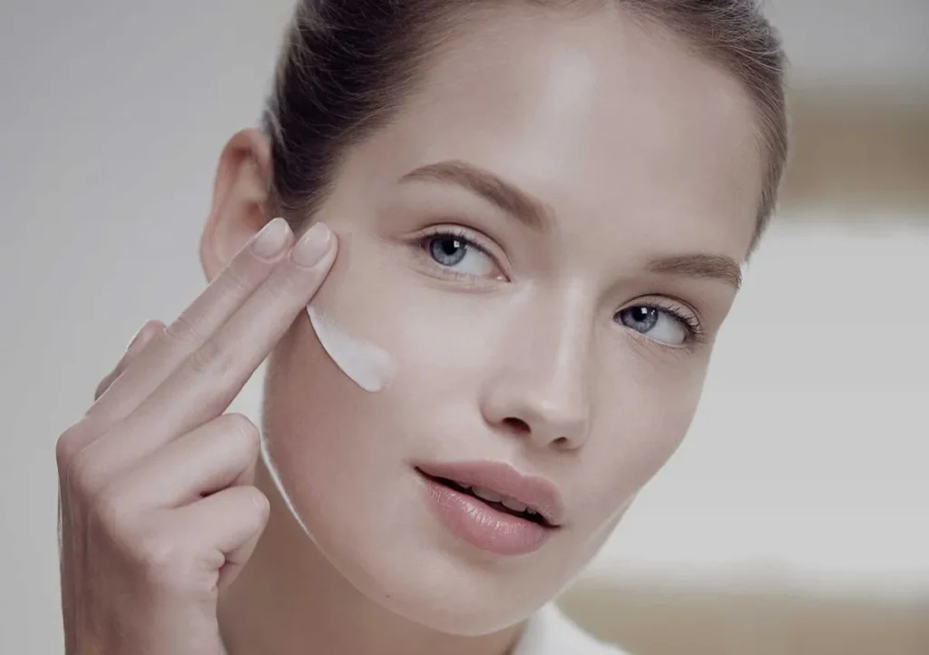 A woman applies a cream to her cheek with her fingers. She has a calm expression, clear skin, and an elegant, minimalist background. Skincare routine.