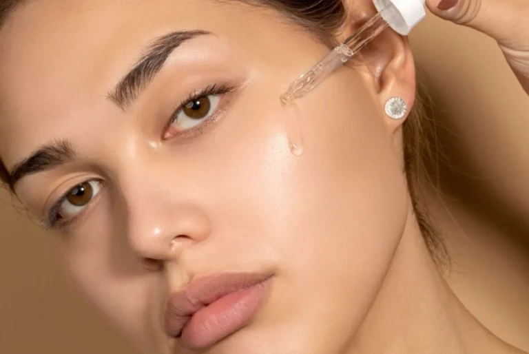 A woman with clear skin applies serum to her cheek using a dropper. Her expression is serene, highlighting a skincare routine. The background is neutral.