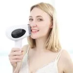 A woman with blonde hair smiles while holding a white hair dryer close to her face. She is wearing a white tank top in a bright, softly lit room.